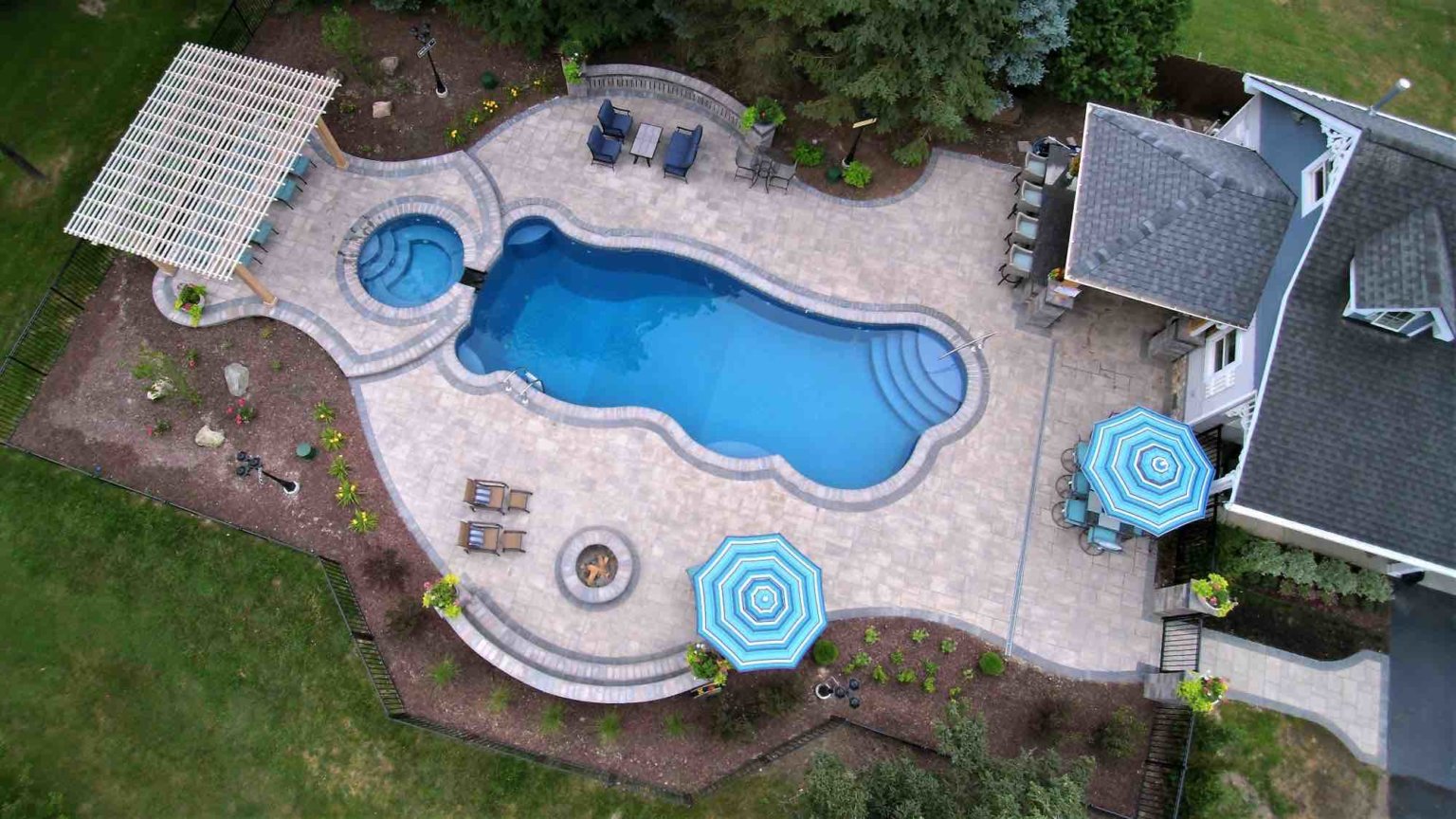aerial view of pool and hot tub with poolside lounge, deck seating, fire pit, and outdoor kitchen